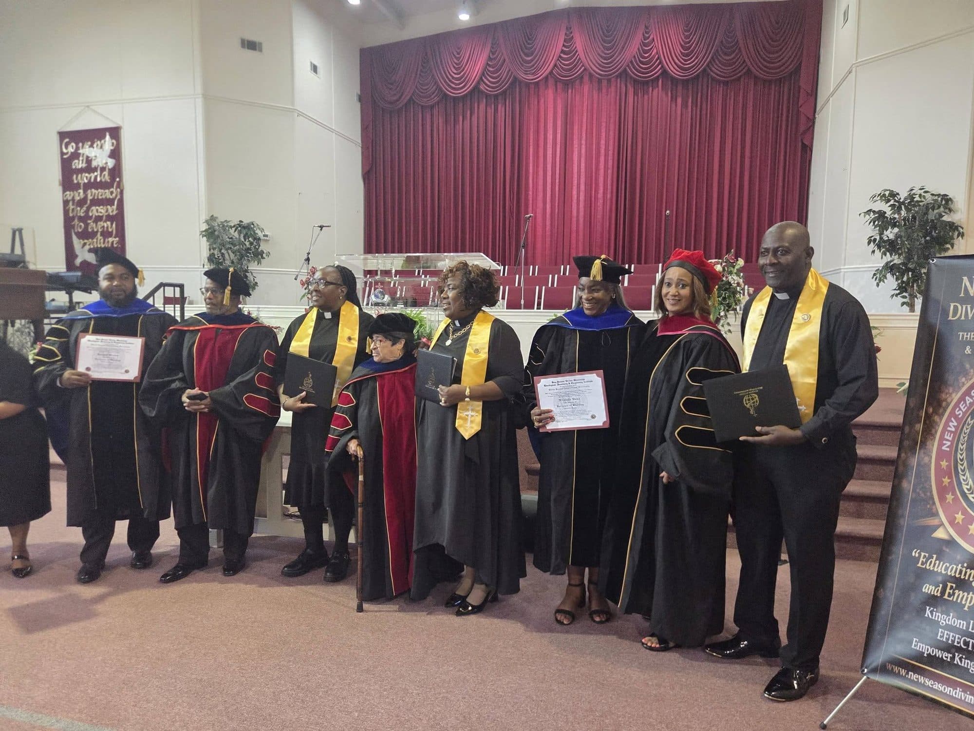 Graduates of the New Season Divine Ministries seminary in doctoral robes and gold chaplain stoles, standing before a red stage curtain with their diplomas