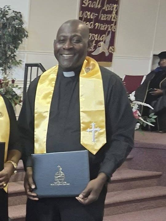 Pastor Heldin C. Gordon smiling in a clerical collar and gold chaplain stole, holding his diploma