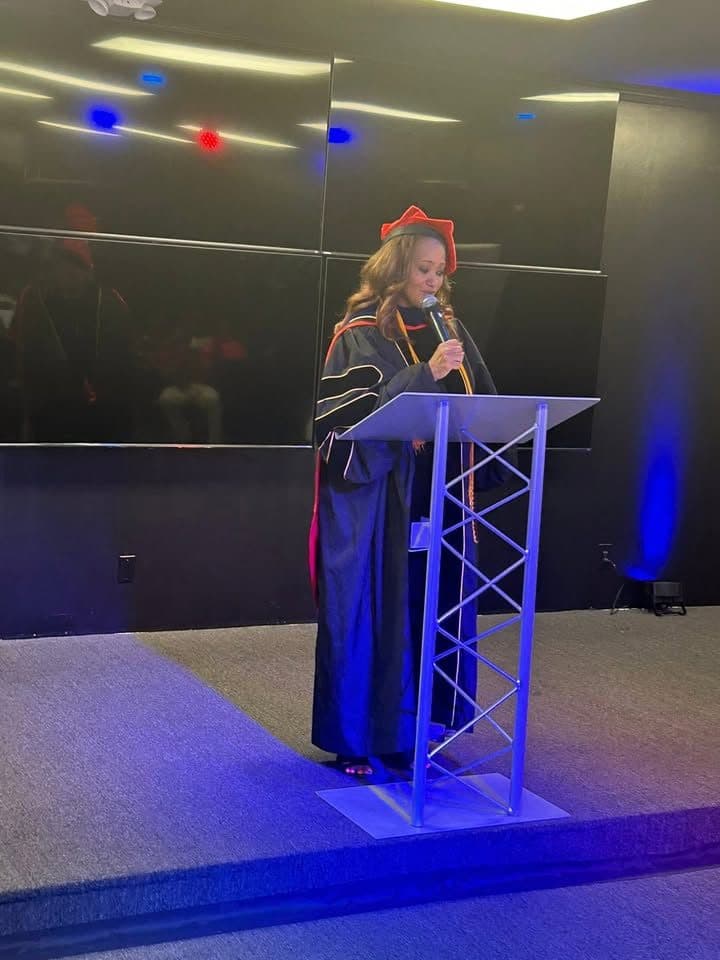 Dr. Sctonda Kelly Gordon in a doctoral robe and red tam, speaking at a podium under blue stage light
